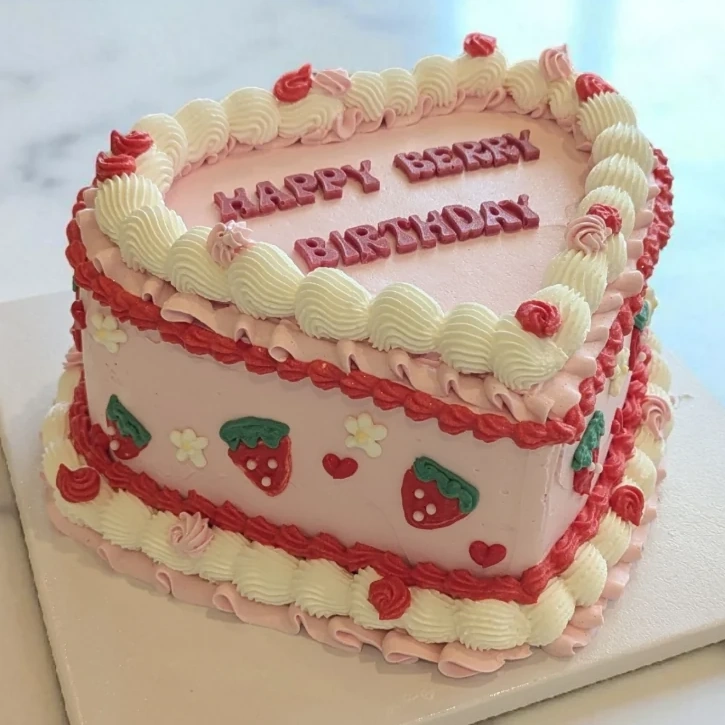 A heart-shaped cake in pale pink buttercream, edged with cream shell piping and red scrollwork, decorated with piped strawberries, daisies and small hearts. "Happy Berry Birthday" is lettered across the top.