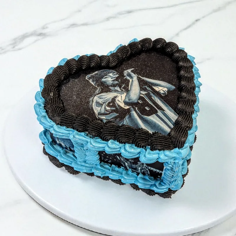 A heart-shaped cake with a printed photographic panel of a singer on stage across the top, framed by a dark chocolate rope border and a bright blue shell border around the sides.