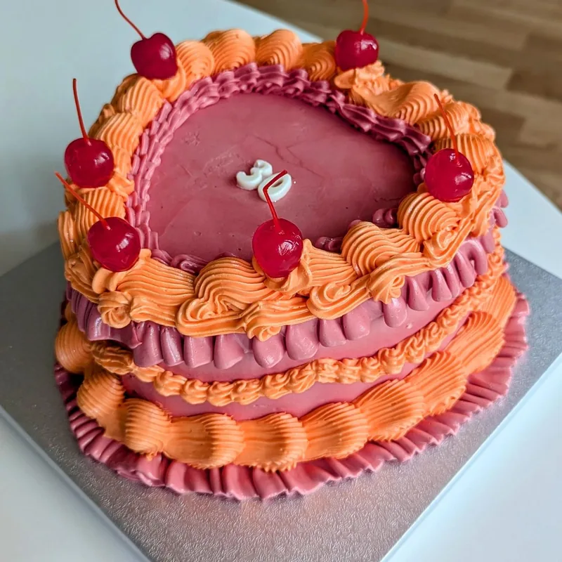 A heart-shaped vintage cake piped in rose pink and orange buttercream, ringed with ruffles and shell borders and topped with glace cherries. A small number three candle stands in the centre.