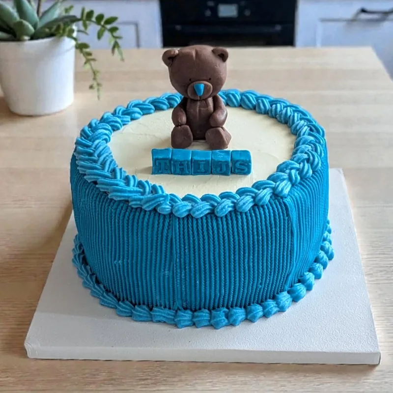 A cake in bright blue buttercream with combed sides and rope borders, its top left cream. A moulded chocolate teddy bear sits on top behind blue fondant letter blocks spelling a name.