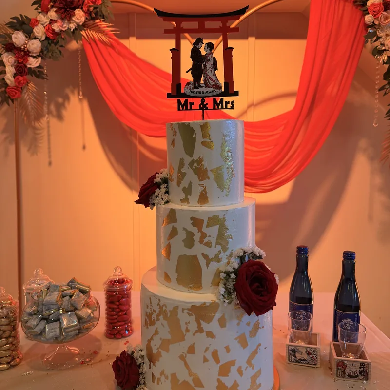 A three-tier wedding cake in white buttercream, leafed with irregular patches of gold and dressed with deep red roses and sprigs of gypsophila. A black acrylic topper of a couple under a torii gate reads "Mr and Mrs".